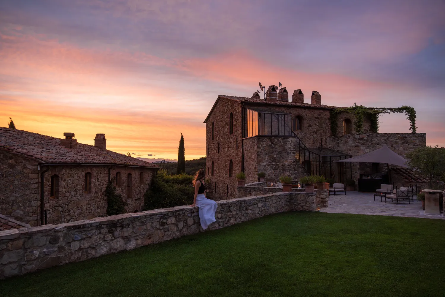 Woman in white dress sits on a stone wall overlooking a Tuscan castle wedding venue, Castello di Vicarello, at sunset.