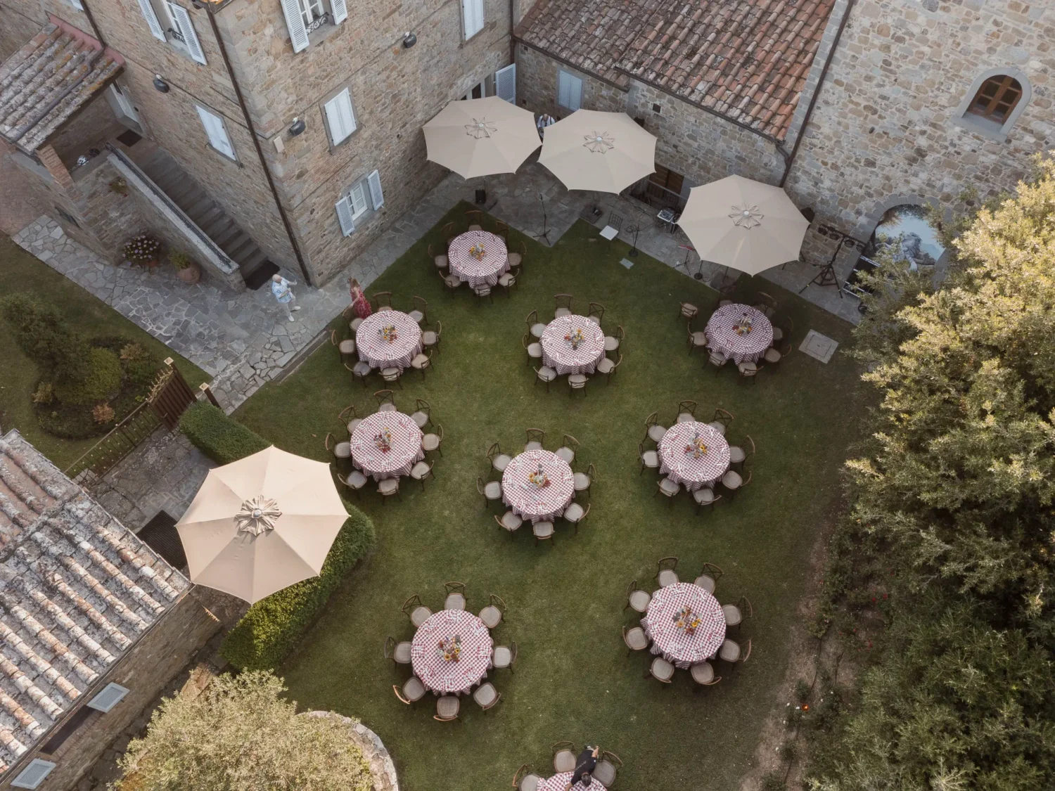 Aerial view of an Italian countryside wedding reception. Tables with red and white checkered tablecloths are arranged on a manicured lawn.