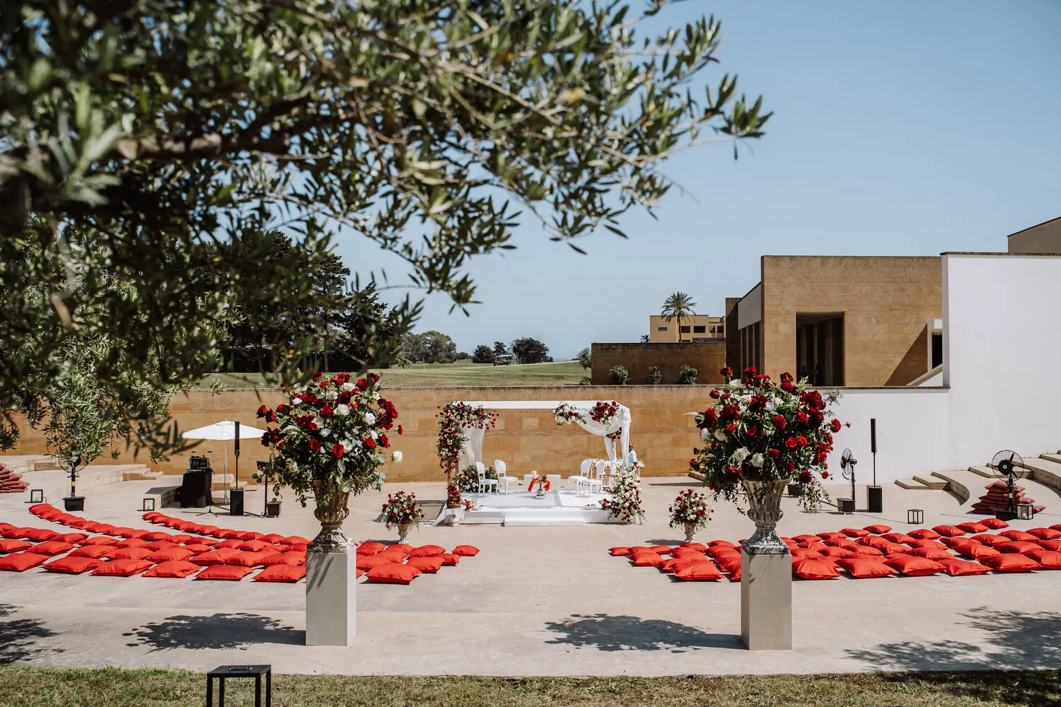 Outdoor Sicilian wedding ceremony setup at Verdura Resort. Red floral arrangements and red cushions flank a white wedding mandap.