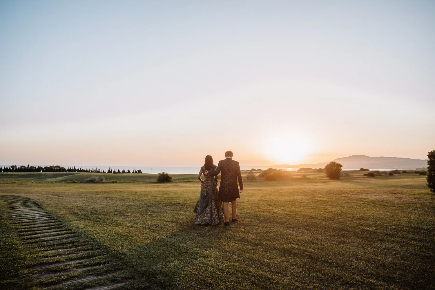 Couple in traditional Indian wedding attire walking away on a Sicilian golf course at sunset. Sicily destination wedding.