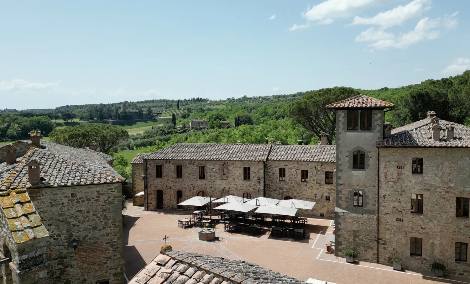Tuscan courtyard at Castel Monastero, perfect for weddings & honeymoons. Stone buildings surround a patio with tables and umbrellas.