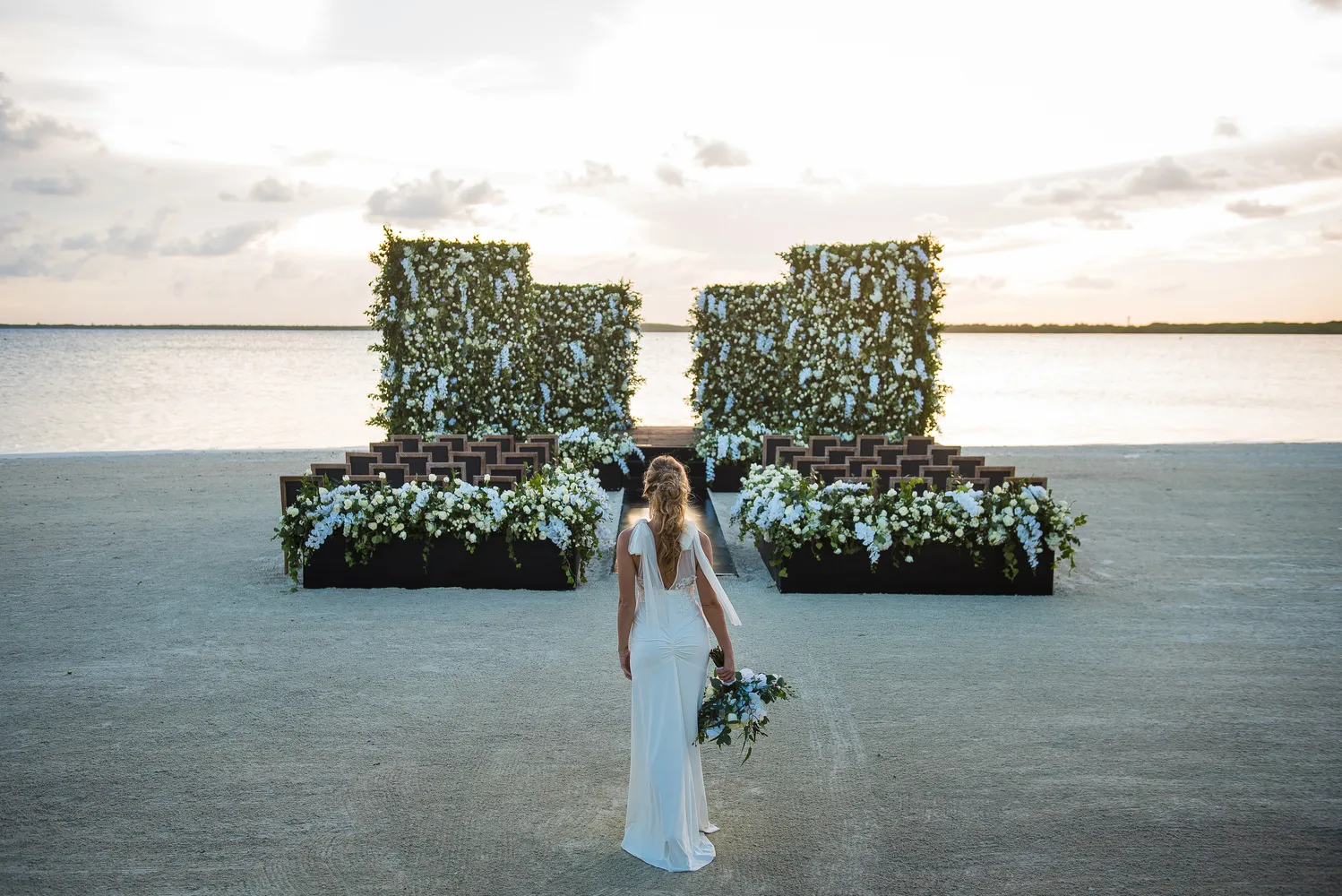 Bride walking toward a beach wedding ceremony at NIZUC Resort & Spa in Cancun. Floral arrangements and seating create a stunning backdrop.