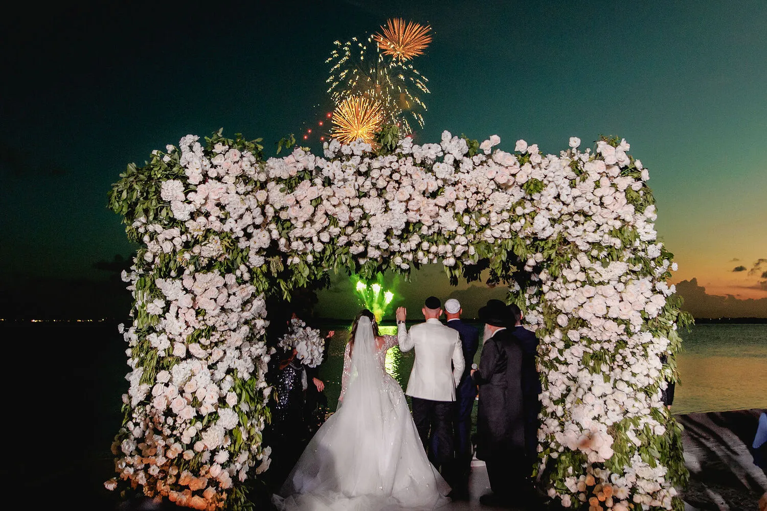 Bride and groom at a Cancun wedding ceremony under a floral archway with fireworks.