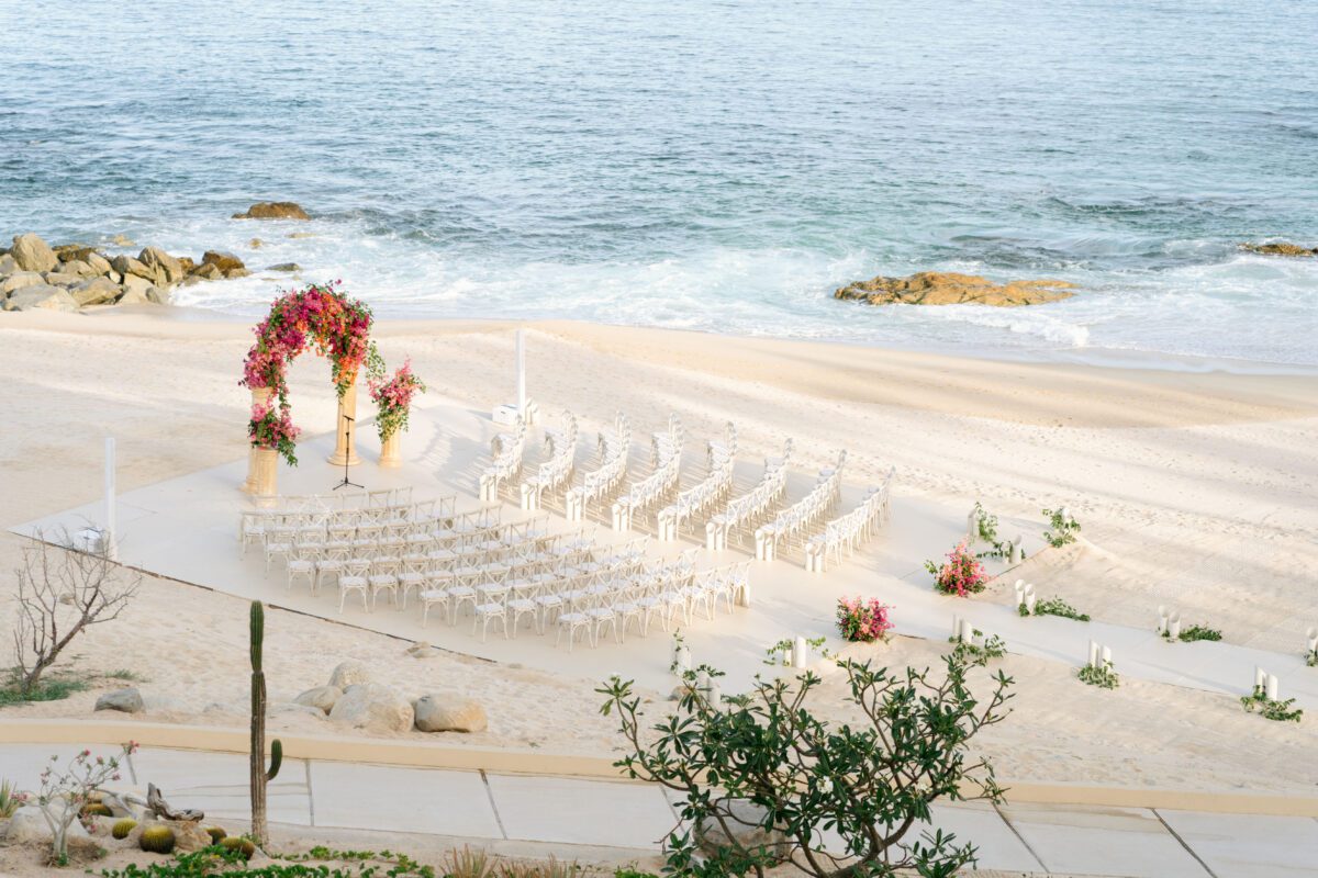 Los Cabos destination wedding setup on a beach, featuring floral arch and rows of white chairs.