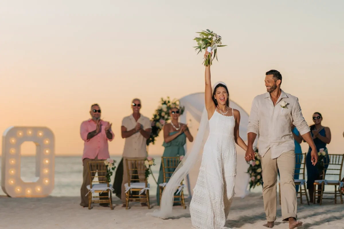 Newlyweds celebrate after a Hilton Aruba Destination Wedding on the beach with guests applauding.