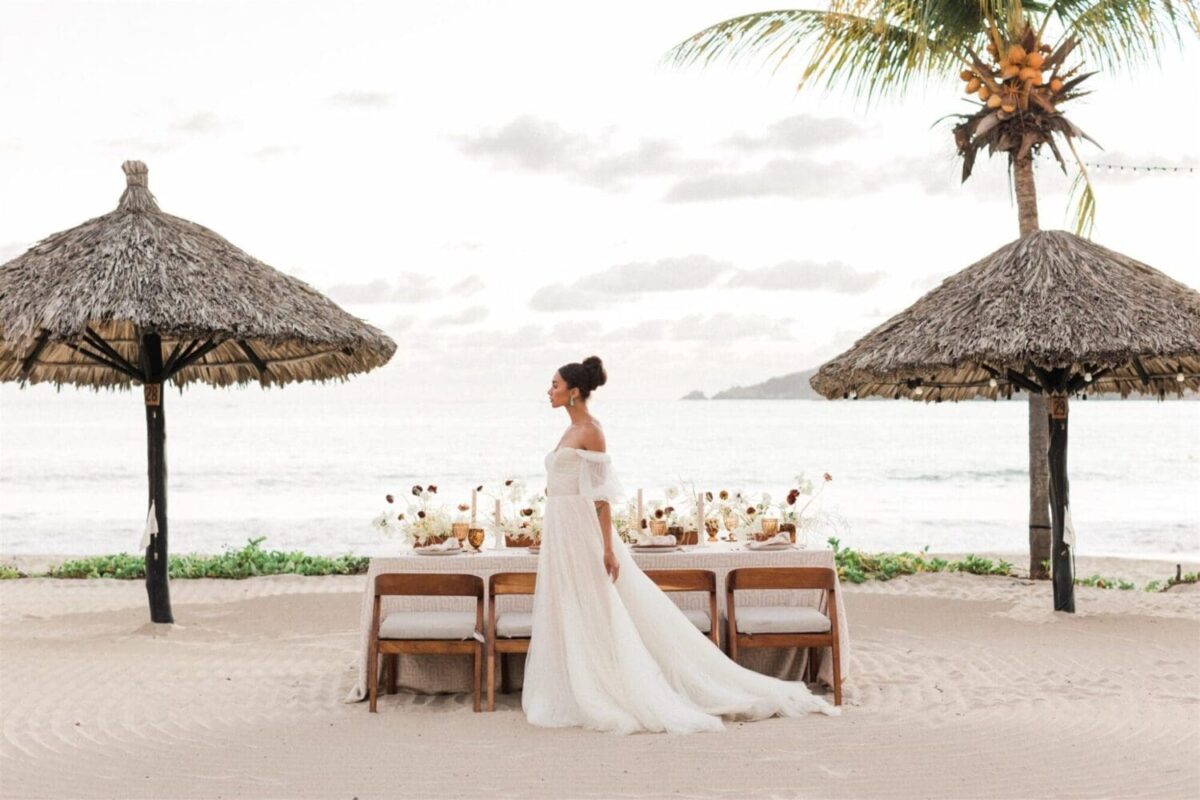 Bride in gown near beachside wedding setup with thatched umbrellas