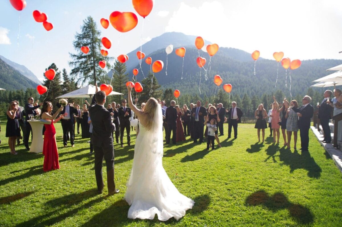 Bride and groom releasing red balloons at outdoor wedding, guests watching