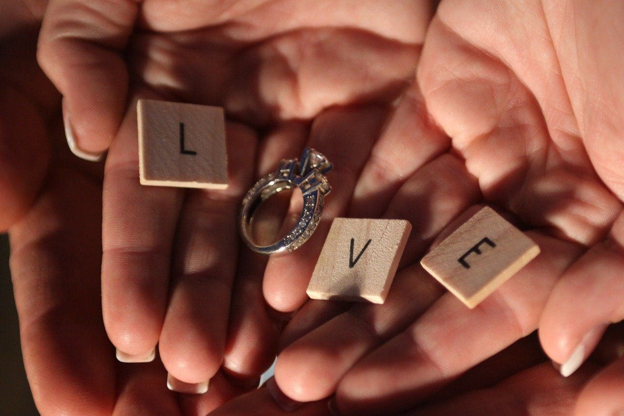 Hands holding Scrabble tiles spelling "LOVE" with an engagement ring