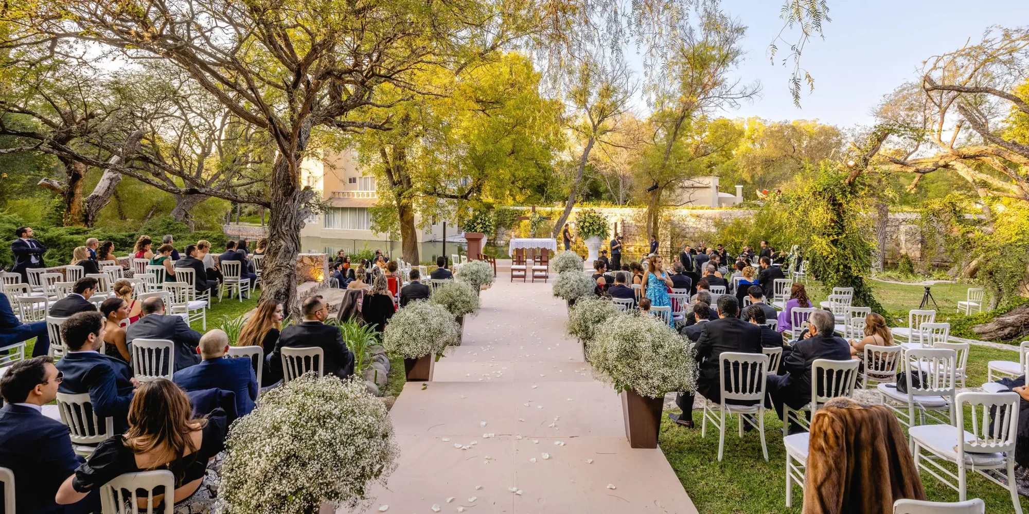 Outdoor wedding ceremony at Agua Bendita in Mexico. Guests seated in white chairs, aisle lined with flowers.