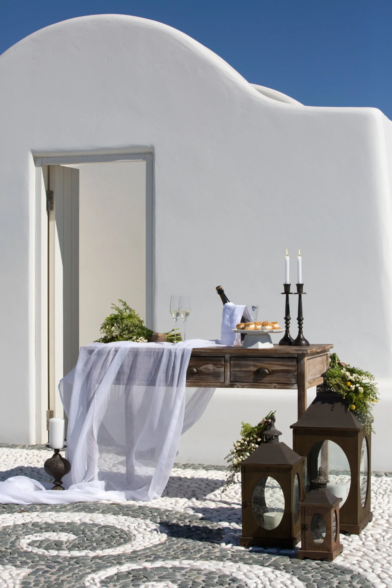 Romantic Santorini wedding setup: wooden table with champagne, pastries, and candles, adorned with flowers and sheer fabric. Dream wedding in Santorini.