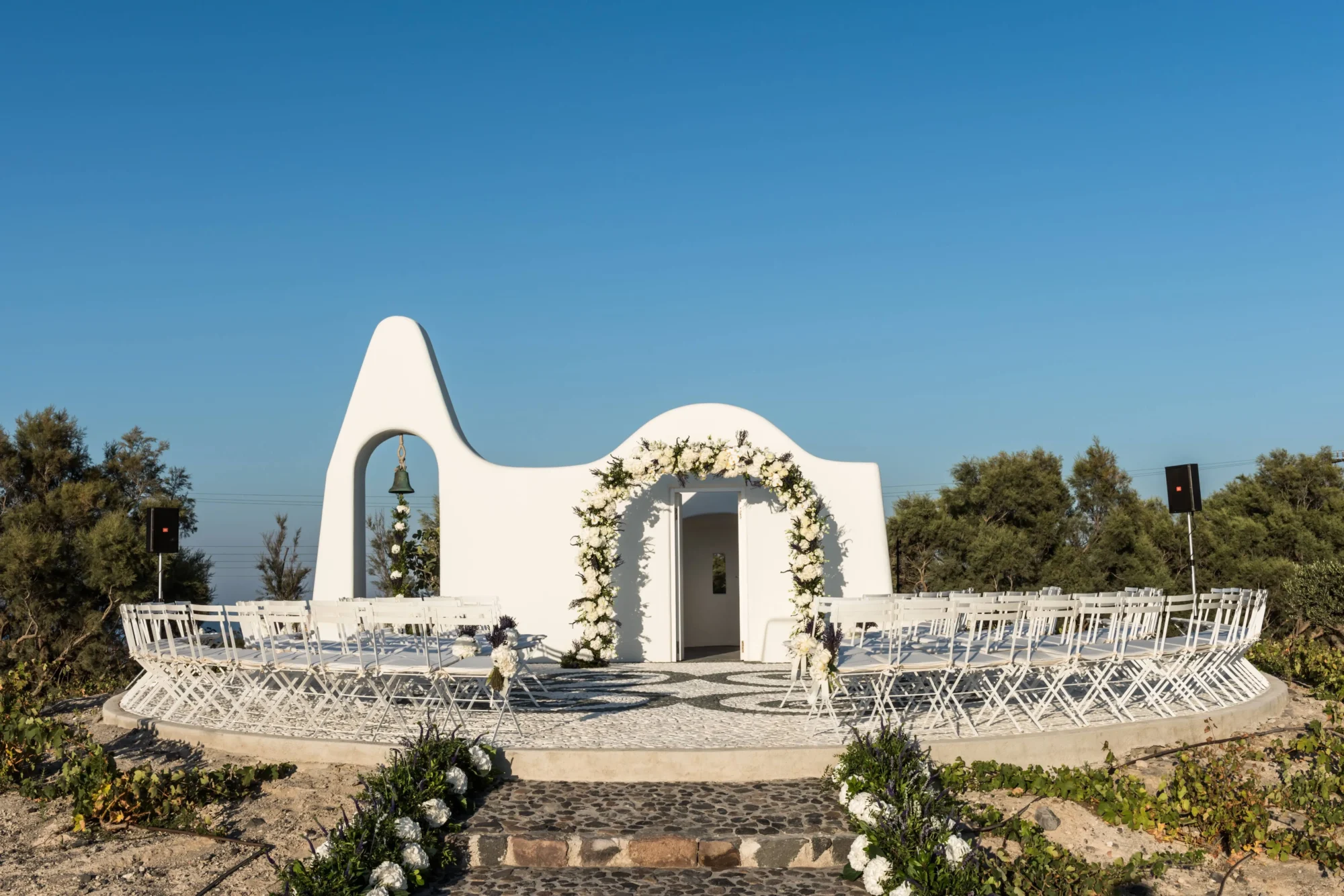 Santorini dream wedding ceremony setup: white chairs, floral arch, and stunning blue sky.