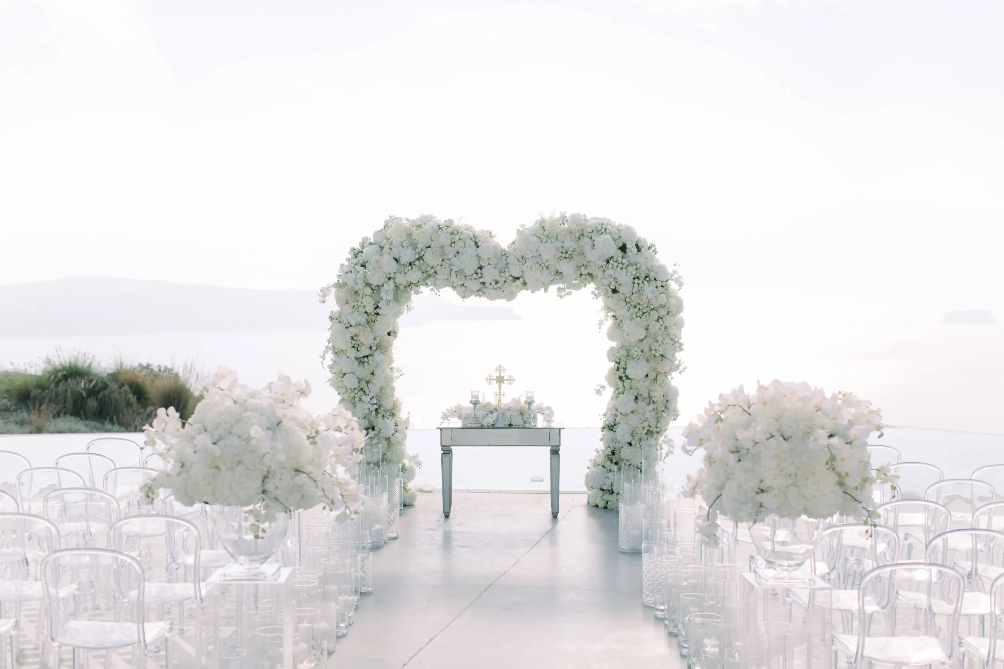 Dream Santorini wedding ceremony with white floral heart arch and clear chairs.