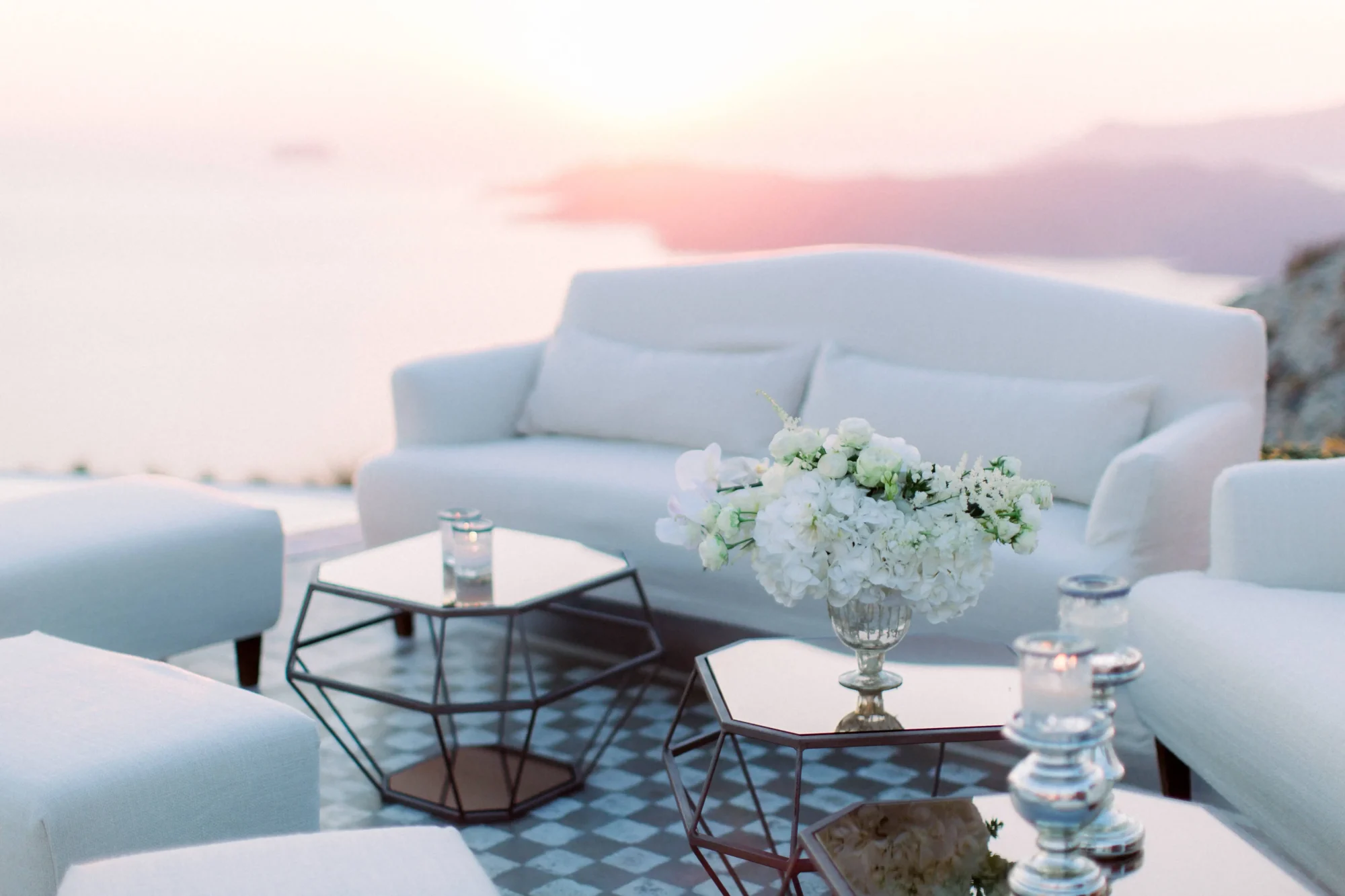 White sofas and geometric coffee tables with white floral arrangement, overlooking Santorini sunset. Dream Santorini wedding.
