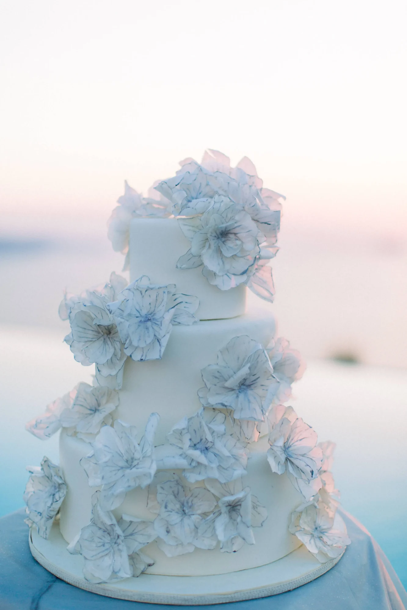 Three-tiered wedding cake adorned with delicate, light blue sugar flowers. Dream Santorini wedding.