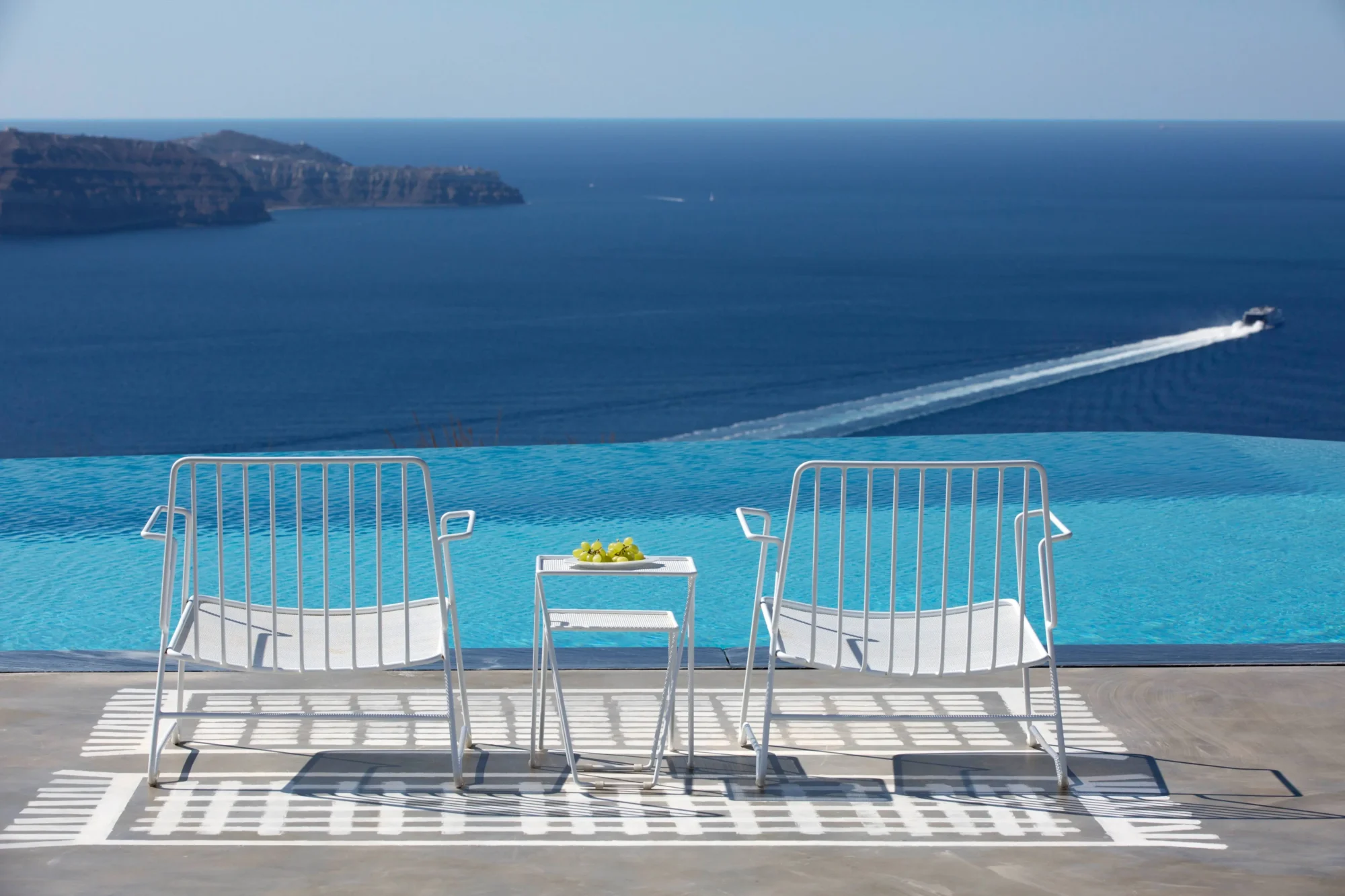 Two white chairs and small table overlooking Santorini's infinity pool; dream wedding location.