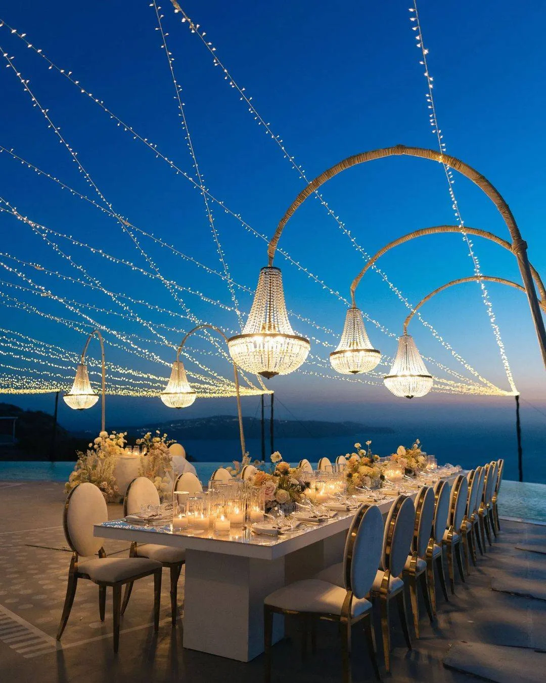 Romantic Santorini wedding reception with a long table, elegant chairs, and crystal chandeliers under a starry sky.