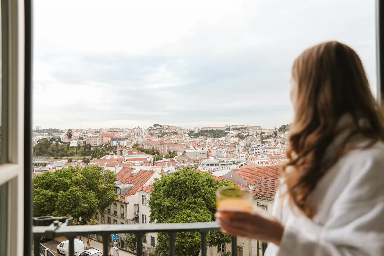 Woman in bathrobe enjoys a drink on a balcony overlooking Lisbon, Portugal.
