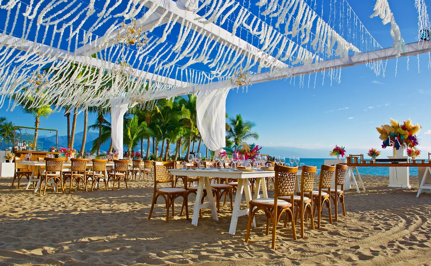 Beachfront destination wedding setup at Marriott Puerto Vallarta. Tables, chairs, and floral arrangements are visible under a white pergola.