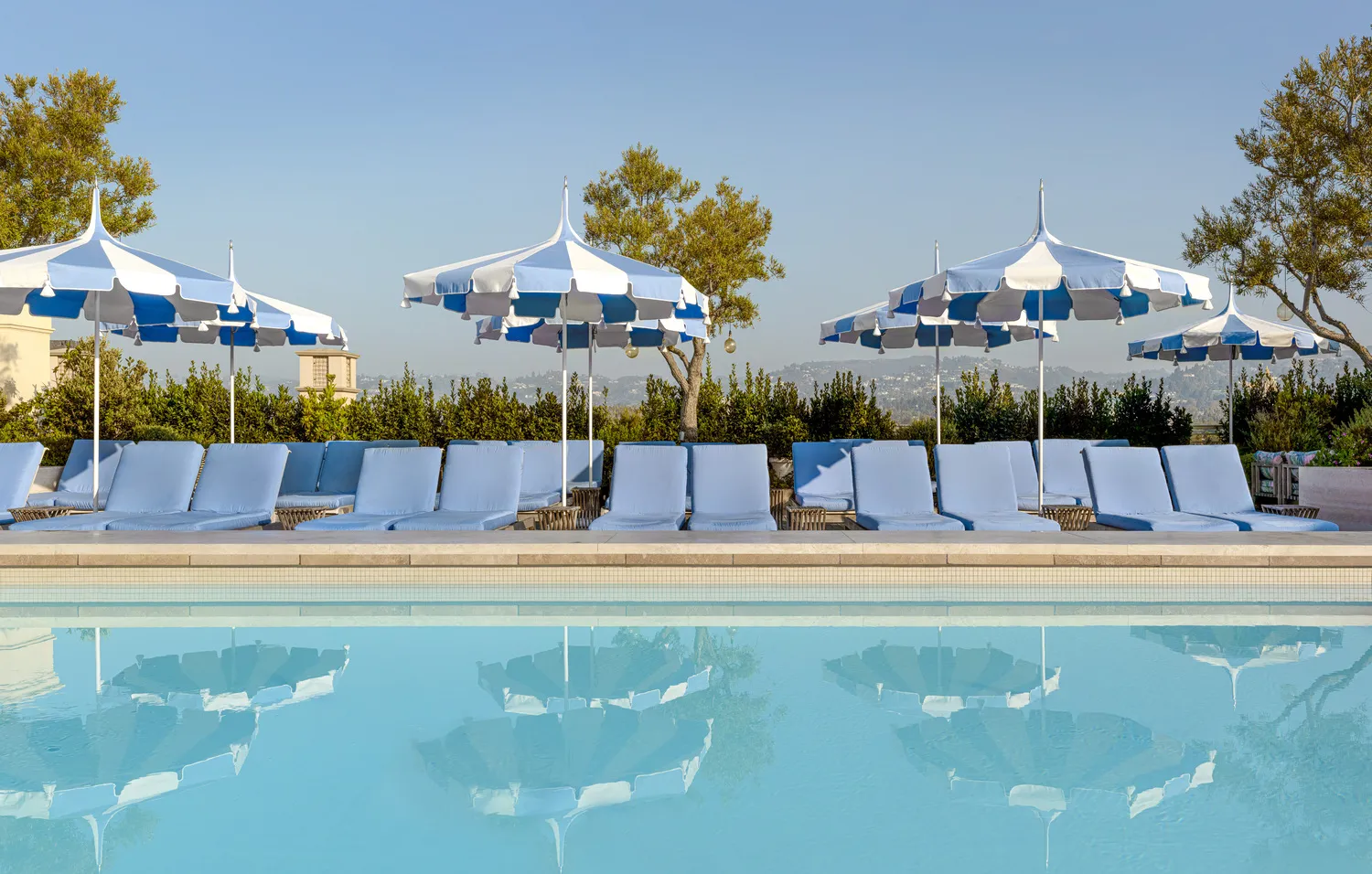 Luxury poolside at Maybourne Beverly Hills, featuring blue lounge chairs and striped umbrellas.