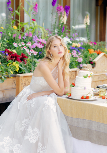 Bride in lace gown seated by a floral wedding cake, surrounded by colorful garden blooms